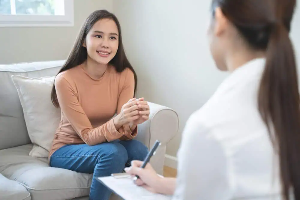 A young woman in a brown sweater and jeans sits comfortably on a beige couch, smiling and conversing with another person who has their back to the camera. The second person appears to be taking notes during what could be a session of Family Therapy in Suffolk County.