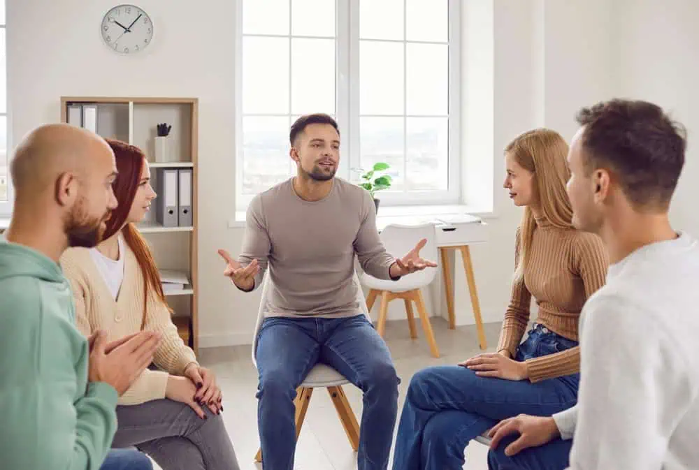 A group of five people sits in a circle in a bright room with large windows. One man, seated in the center, is speaking and gesturing with his hands, while the others listen attentively. The setting appears to be a psychotherapy session, possibly Couples Therapy Suffolk County.