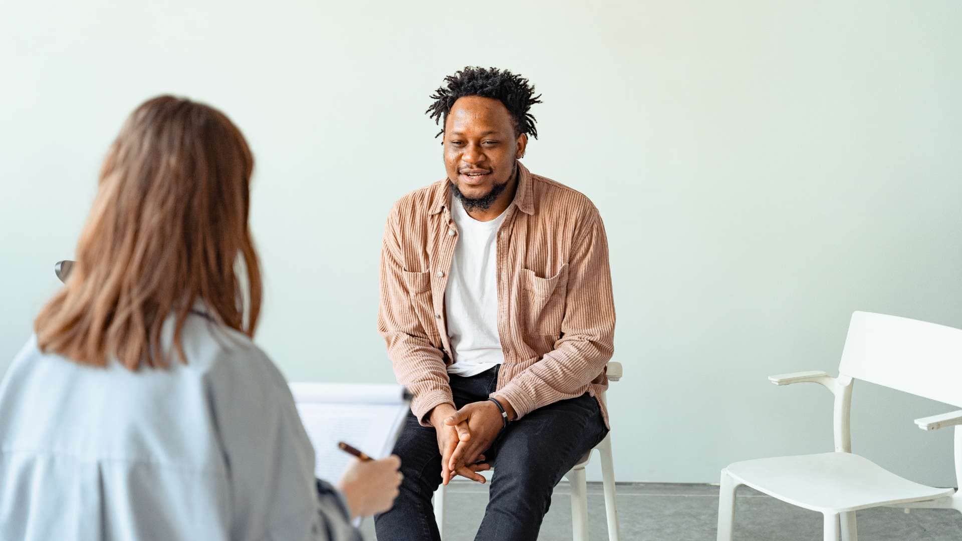 In a bright, minimalist room with white walls and white chairs, a man with earphones sits and smiles while talking to a person with long hair. The man, clad in a brown jacket and dark pants, speaks animatedly as the other person holds a pen and notebook, capturing the essence of Couples Therapy Suffolk County.