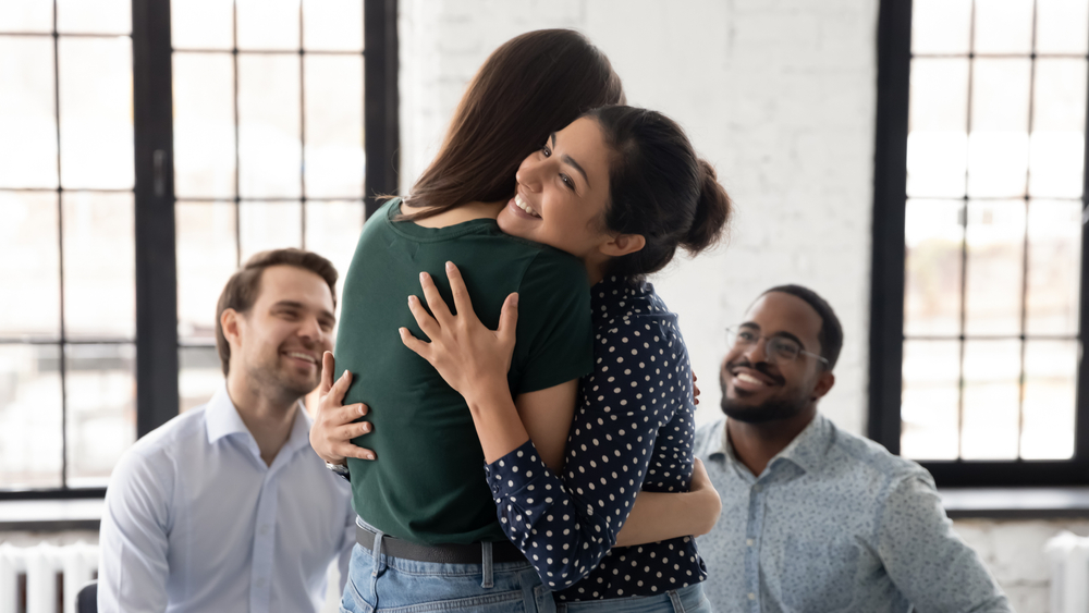 A group of four people in a room, featuring two women hugging each other warmly. Two men sit nearby, watching the pair with happy expressions. The setting suggests a supportive and friendly atmosphere.