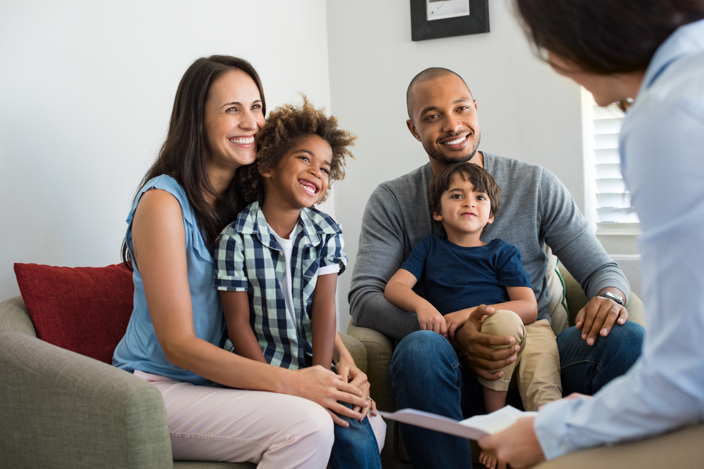 A happy family of four sits on a couch, smiling and listening to a person off-frame. Two adults sit with two young children, one boy and one girl. The setting appears to be a comfortable meeting or living room.