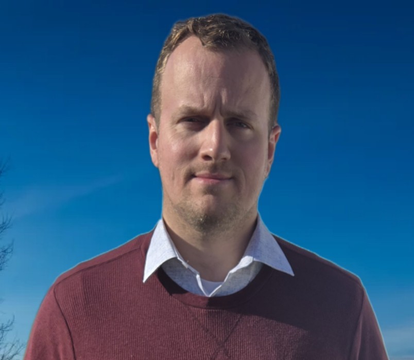 Under the clear blue sky, a person with short hair stands wearing a maroon sweater over a white collared shirt, embodying the essence of our About Us story—a neutral expression reflecting simplicity and authenticity.