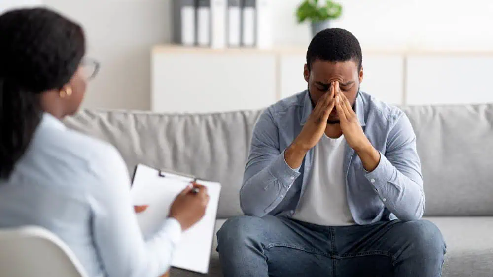 A man sits on a couch, leaning forward with his hands pressed together against his forehead, appearing stressed. He faces a seated person holding a clipboard, suggesting a therapy or counseling session in a comfortable room setting.
