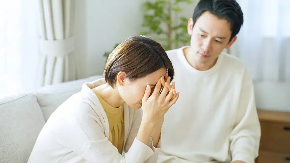 A woman sitting on a couch, holding her head in her hands, appears distressed. A man next to her looks concerned and supportive. They are in a well-lit room with white curtains and a green plant in the background.