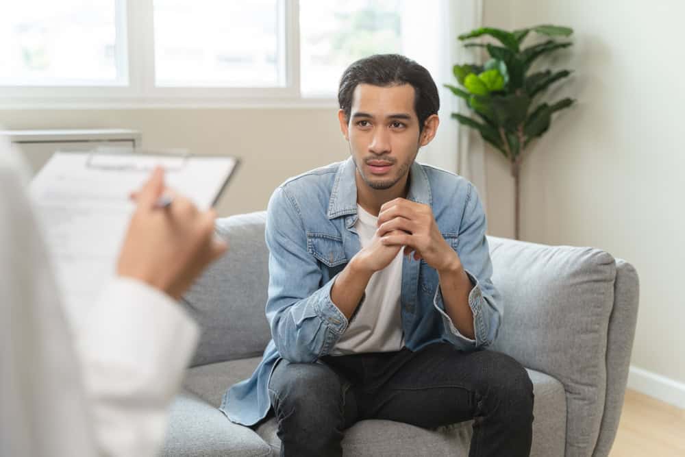 A man sits on a gray sofa, attentively listening to someone holding a clipboard. He wears a denim jacket and black jeans. A houseplant is in the background, and soft daylight comes through a window.