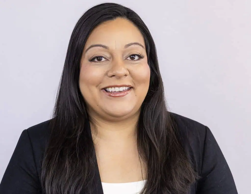 A woman with long dark hair, wearing a black blazer over a white top, smiles at the camera against a plain light gray background—perfect for an About Us introduction.