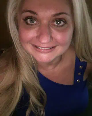 About Us: A woman with long blonde hair and blue eyes smiles at the camera, wearing a sleeveless royal blue top with gold buttons on one shoulder.