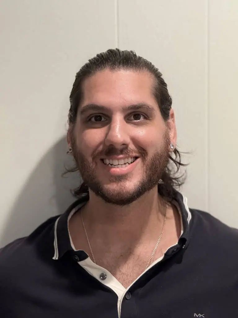 A head-and-shoulders portrait of a man with long, dark hair tied back. He has a full beard and is smiling directly at the camera. He is wearing a dark polo shirt with a white collar.