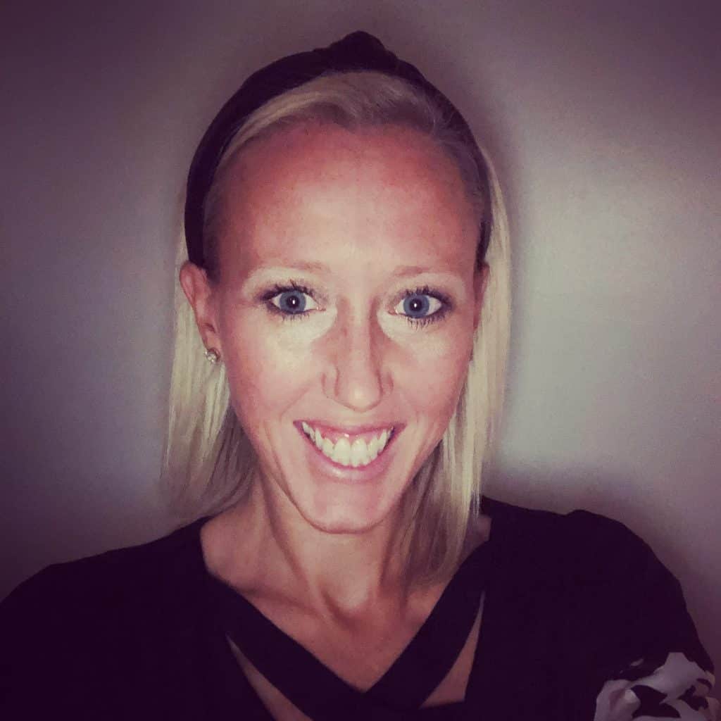 A woman with straight blonde hair, wearing a black headband and a black top, smiles at the camera against a plain background. The soft lighting and vignette effect reflect the welcoming spirit of our About Us page.