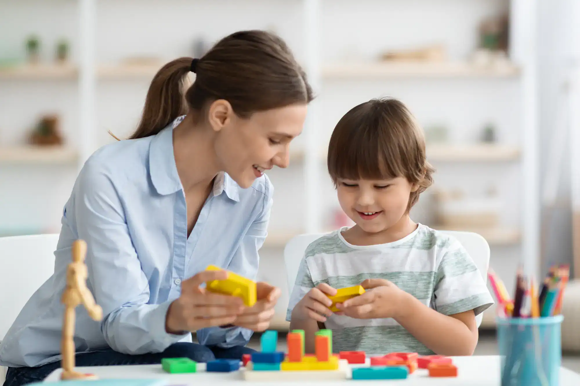 A woman and a young child sit at a table, smiling and playing with colorful wooden blocks together in a bright room.